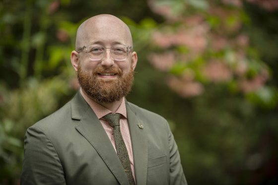 person in an olive green suit and clear glasses looking at the camera